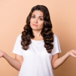 Photo of dislike young curly hairdo lady shrug shoulders wear white t-shirt isolated on beige color background.